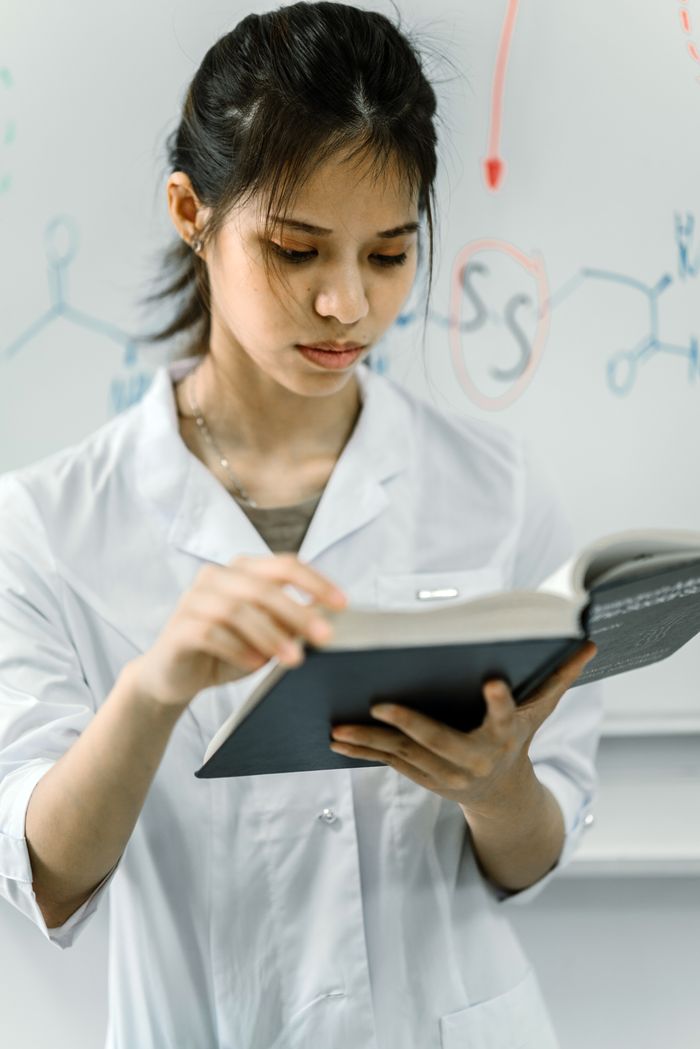 services-03 Scientist in lab coat reading a book in front of a chemistry-themed whiteboard.